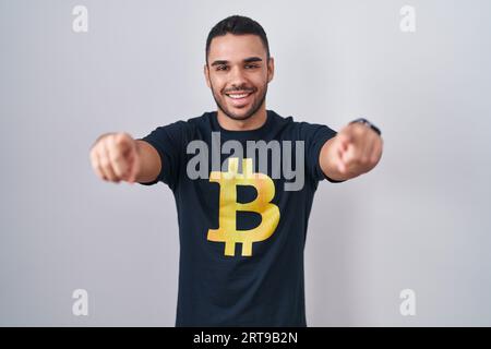 Jeune homme hispanique portant un t-shirt bitcoin pointant vers vous et la caméra avec les doigts, souriant positif et joyeux Banque D'Images