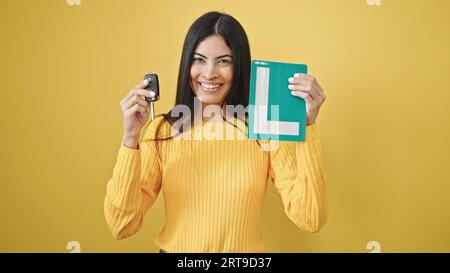 Jeune belle femme hispanique tenant la clé de la nouvelle voiture et le nouveau permis de conduire sur fond jaune isolé Banque D'Images