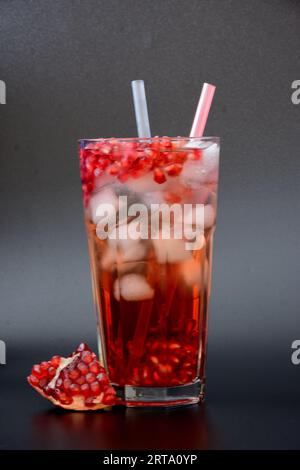 Un grand verre à facettes de jus de grenade rafraîchissant avec de la glace et des tubes sur un fond noir, à côté se trouve un fruit mûr cassé avec des graines. Vertical Banque D'Images