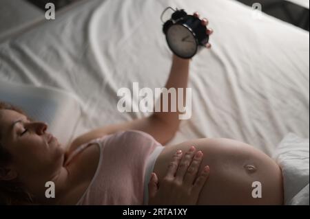 Une femme enceinte se couche dans son lit et tient un réveil. Banque D'Images