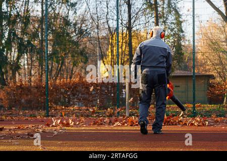 Homme aux cheveux gris portant des cache-oreilles à l'aide d'un souffleur de feuilles portable pour nettoyer la zone clôturée du parc. Vue arrière de l'homme senior en salopette bleu foncé, soufflant les feuilles du terrain de jeu. Concept de travail. Banque D'Images