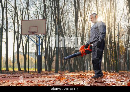 Concierge souriant avec le nettoyage des cheveux gris parc de la ville d'automne avec souffleur de feuilles. Vue latérale d'un homme âgé portant des vêtements de travail spéciaux et des cache-oreilles remov Banque D'Images