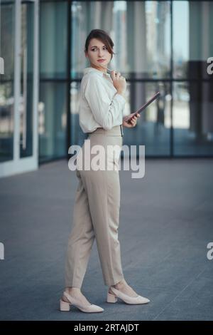 Jeune femme d'affaires mignonne caucasienne dans le style de bureau Pantsuit dans les tons beiges avec tablette numérique dans les mains posant près de l'immeuble de bureaux Banque D'Images