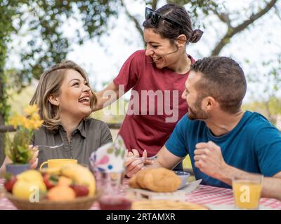 Trois amis caucasiens profitent d’un brunch en plein air. Une femme assise et un jeune homme ont une étendue de nourriture devant eux, tandis qu'une femme brune se tient debout, embr Banque D'Images