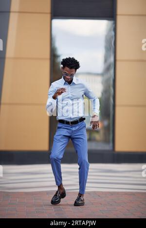 Heureux homme noir dansant sur la rue de la ville près du bâtiment, souriant et portant des vêtements formels pantalons bleus et chemise. Beau modèle masculin à l'extérieur. Banque D'Images