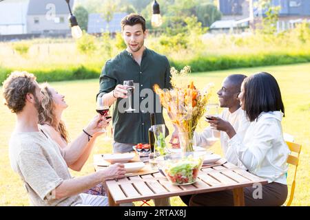 Un moment délicieux lors d'une Garden party où un groupe diversifié d'amis millénaires est assis autour d'une table à dîner magnifiquement fixée. Au milieu d'une conversation joyeuse et de rires, un charismatique homme d'Europe du Sud se lève, tenant son verre de vin haut. Il lui propose un toast du cœur, et à l’unisson, les autres le rejoignent en levant leurs verres, en célébrant l’amitié et les bons moments. Des amis milléniaux multiraciaux lèvent des lunettes en toast. Photo de haute qualité Banque D'Images