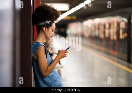 Vue latérale de jeune femme marocaine avec bandeau de cheveux afro regardant l'écran du téléphone portable tout en se tenant debout vérifier les messages texte sur la plate-forme du métro Banque D'Images