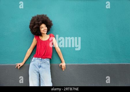 Souriant mignon jeune femme marocaine avec coiffure afro regardant loin tout en se tenant debout appuyé sur le mur bleu avec les mains sur la surface grise dans la lumière du jour Banque D'Images