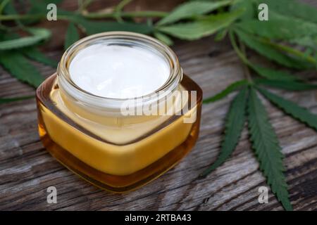 Crème hydratante en pot avec des feuilles de chanvre sur fond en bois. Crème cosmétique pour les soins de la peau à partir de chanvre. Cosmétiques naturels Banque D'Images