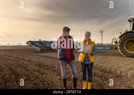 Homme et femme fermiers matures debout dans le champ, parlant sur mobile et écrivant des notes tout en labourant le tracteur en arrière-plan Banque D'Images