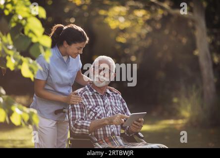 Homme senior en fauteuil roulant montrant quelque chose sur tablette à la jeune infirmière soignante derrière lui dans le parc Banque D'Images