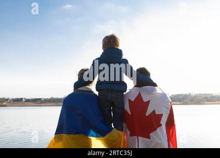 Femme avec drapeau ukrainien, homme avec drapeau canadien derrière le dos et enfant , assis dos ensemble dans la nature . Appui et assistance internationaux Banque D'Images