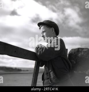 Années 1950, historique, un garçon berger appuyé sur une porte rurale en bois regardant à travers un champ, Angleterre, Royaume-Uni. Banque D'Images