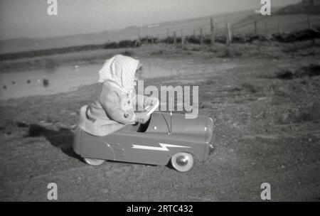 Années 1950, historique, à l'extérieur sur un terrain dégagé, dans une zone herbeuse près d'un étang naturel, une petite fille portant un manteau et un foulard, conduisant / jouant dans une voiture de jouet, Angleterre, Royaume-Uni. Banque D'Images