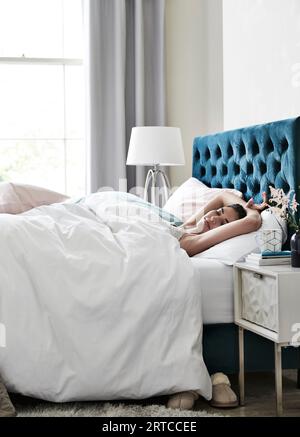 Beau jeune modèle préparant pour le lit, dormir dans le lit scène de chambre avec literie et rideaux avec fenêtre, couette, oreillers style de vie, sereine détendue Banque D'Images