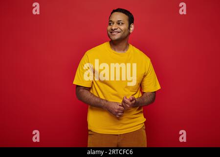 charmant homme indien dans des vêtements décontractés lumineux regardant loin et souriant sur fond rouge dans le studio Banque D'Images