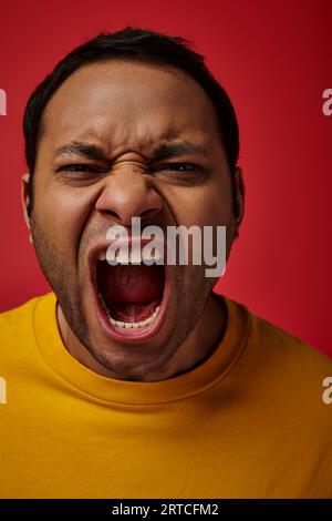 expression du visage, homme indien émotionnel en t-shirt jaune criant sur fond rouge, bouche ouverte Banque D'Images