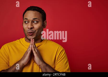 homme indien rusé en t-shirt jaune serrant les mains et planifiant quelque chose sur fond rouge, visage rusé Banque D'Images