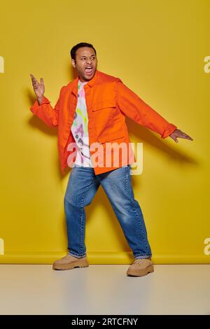 homme indien émotionnel en veste orange et jeans faisant des gestes et criant sur fond jaune Banque D'Images