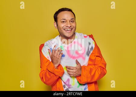 homme indien joyeux en veste orange et t-shirt diy souriant et regardant la caméra sur fond jaune Banque D'Images