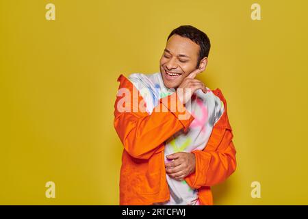 homme indien joyeux en veste orange et t-shirt diy souriant avec les yeux fermés sur fond jaune Banque D'Images