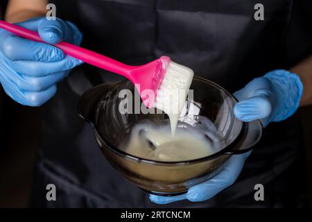 fille dans des gants de protection en caoutchouc bleu tenant une brosse dans ses mains et remuant colorant capillaire gros plan coloriste soins capillaires au masque capillaire à la maison. Banque D'Images