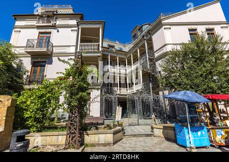 Un bâtiment résidentiel avec des balcons enveloppants peints en blanc se dresse dans la rue Ioane Shavteli dans la vieille ville de Tbilissi, en Géorgie. Des colonnes élancées, des rampes délicates et des portes en fer forgé structurent la cour. À droite, un escalier en colimaçon en fer forgé monte à l'extérieur des balcons. Au premier plan, une plante grimpante aux fleurs rouges pousse sur un treillis métallique. À droite se trouvent un chariot de glaces sous un parasol bleu et un étal avec un toit rouge. Banque D'Images