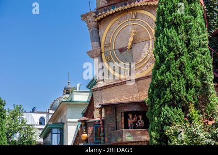 La tour penchée de l'heure du théâtre de marionnettes Rezo Gabriadze au 13 rue Ioane Shavteli dans la vieille ville de Tbilissi, en Géorgie, porte un grand cadran avec des chiffres romains dorés. En dessous, des marionnettes d'un homme et d'une femme se tiennent dans une petite fenêtre. L'homme tient un petit enfant. Deux colonnes empilées avec des chapiteaux soutiennent le coin de la tour. Une maison avec une véranda en bois vert et un bâtiment en dôme se trouvent à gauche. Un cyprès pousse à droite. Chaque heure à l'heure, un ange sort d'une petite porte en haut et frappe la cloche. À 12 heures midi et à 19 heures, un petit théâtre de marionnettes se tient dans la tour Banque D'Images