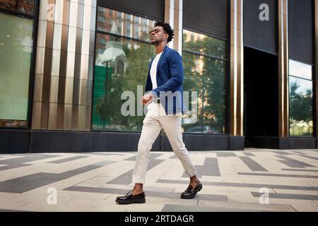 Vue latérale homme pleine longueur marche à l'extérieur et porte des vêtements d'affaires tendance, veste bleue, pantalon blanc et chaussures mocassins. Modèle masculin de mode élégant, Stre Banque D'Images