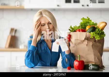 Dépenses d'épicerie. Dame principale stressée dans la cuisine vérifiant la facture après avoir fait des achats de nourriture Banque D'Images