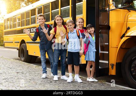 Groupe d'enfants heureux debout près de l'autobus scolaire et faisant signe à la caméra Banque D'Images