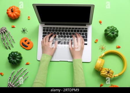 Mains féminines avec ordinateur portable, écouteurs et décorations d'Halloween sur fond vert Banque D'Images