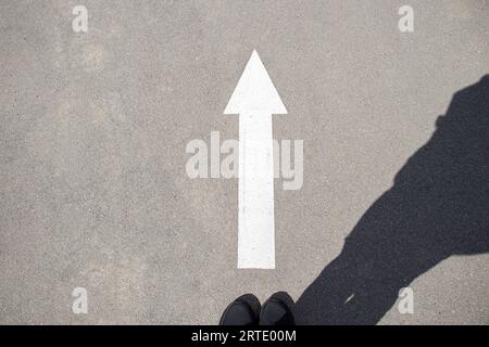 Une femme en baskets se tient près d'une flèche blanche avec une direction vers l'avant sur l'asphalte et une ombre d'une femme, une route avec une flèche vers l'avant Banque D'Images