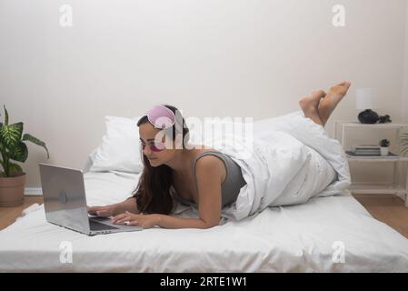 Jeune femme avec masque de sommeil et patchs se trouve sur le lit dans la chambre à coucher regardant l'écran d'ordinateur portable avec le sourire Banque D'Images