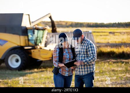 Un couple de fermiers utilisant un appareil portable et sans fil pour gérer et surveiller leur chute, la récolte de canola tout en se tenant devant une moissonneuse-batteuse... Banque D'Images