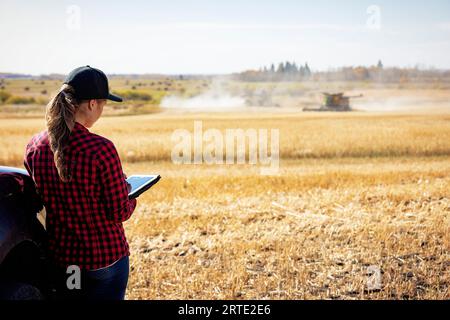 Vue prise de derrière d'une jeune femme agricole debout dans un champ de céréales à côté d'un camion au moment de la récolte, en utilisant le logiciel agricole avancé techno... Banque D'Images