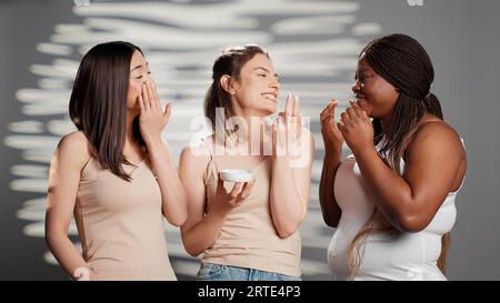 Groupe de belles filles appliquant la crème hydratante sur la caméra, faisant la promotion des produits de soins de la peau naturels en studio. Diverses femmes utilisant hydratant et sérum pour la nouvelle campagne de beauté. Banque D'Images