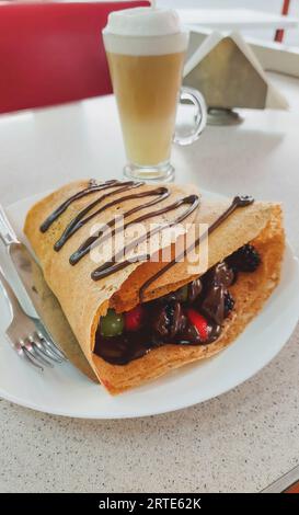 Vue verticale d'une crêpe de fruits rouges avec du chocolat, contenant des fraises, des mûres, des raisins et du chocolat fondu, avec un café cappuccino dans le Banque D'Images