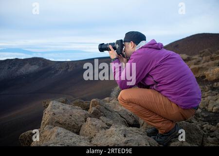 Homme accroupi sur les roches volcaniques sur un sommet de montagne au-dessus des nuages à Haleakala regardant le lever du soleil sur la côte Pacifique et photographiez... Banque D'Images