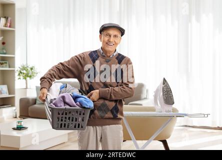 Homme âgé tenant un panier à linge plein de vêtements devant une planche à repasser dans un salon Banque D'Images