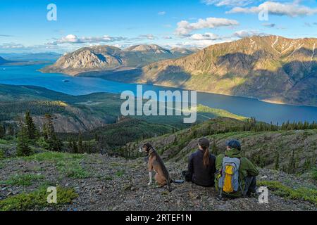 Vue prise de derrière de deux femmes assises au sommet d'une montagne avec leur chien profitant de la vue, tout en explorant et photographiant la scène étonnante... Banque D'Images