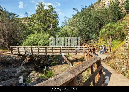 Vila de Rei, Portugal - 20 avril 2023 : passerelles Penedo Furado avec un pont en bois sur le ruisseau et deux personnes assises sur un banc reposant sur un soleil Banque D'Images