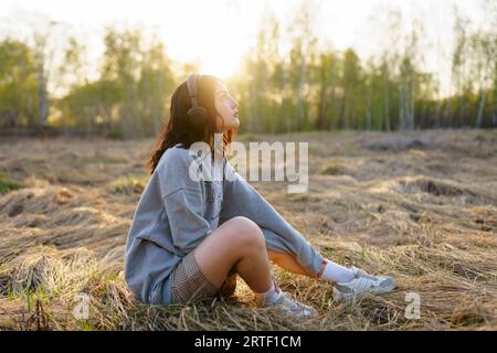 Vue latérale de la femme écoutant de la musique tout en étant assise sur l'herbe dans le pré au coucher du soleil Banque D'Images