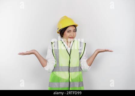 Une jeune femme réfléchie de travail portant casque de sécurité et gilet est confondue entre les choix à côté d'elle, isolé par fond blanc. Concept de fête du travail Banque D'Images