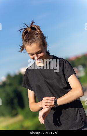 Adolescente en vêtements noirs vérifiant sa montre de fitness après une séance d'entraînement. Fille coureuse fatiguée contrôlant les pulsations vérifiant smartwatch. Banque D'Images