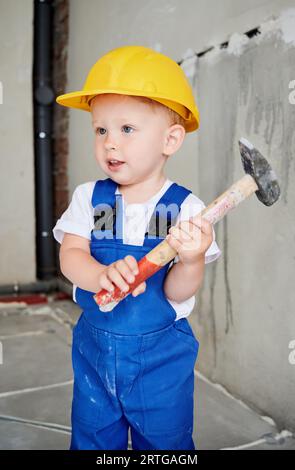 Adorable ouvrier de construction d'enfant tenant l'outil de réparation de construction de marteau. Enfant mignon portant un casque de sécurité et une combinaison de travail tout en posant à la maison pendant la rénovation. Banque D'Images