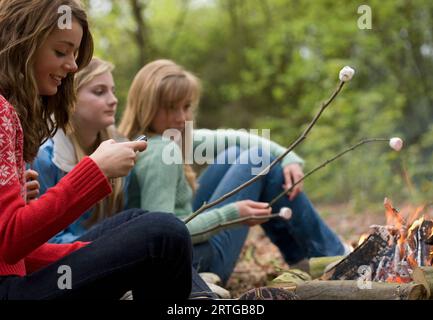 Des adolescentes rôtissant de la guimauve sur un feu de camp on utilise un téléphone portable Banque D'Images