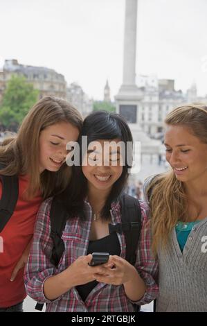 Sourire trois adolescentes en utilisant un téléphone cellulaire à Londres Trafalgar Square Banque D'Images