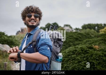 homme indien joyeux dans les lunettes de soleil et chemise en denim vérifiant l'heure sur la montre-bracelet, randonneur avec l'équipement de voyage Banque D'Images