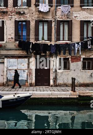 Homme en noir marchant le long du canal vénitien, ancienne maison historique derrière avec des lignes de lavage accrochant des vêtements pour sécher. Bateau en premier plan Venise, Italie Banque D'Images